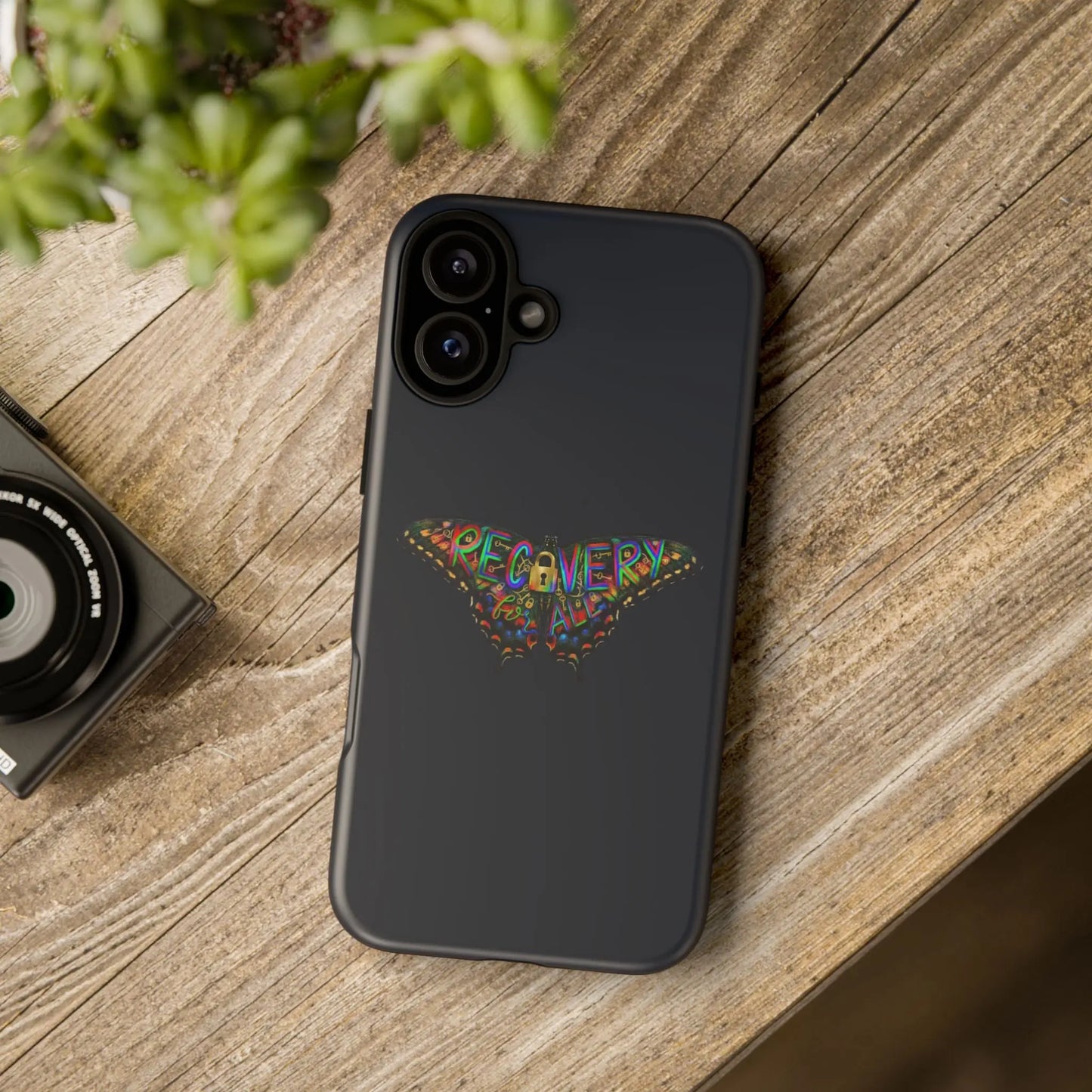 Phone case with motivational quote on a wooden surface with a camera and plant.