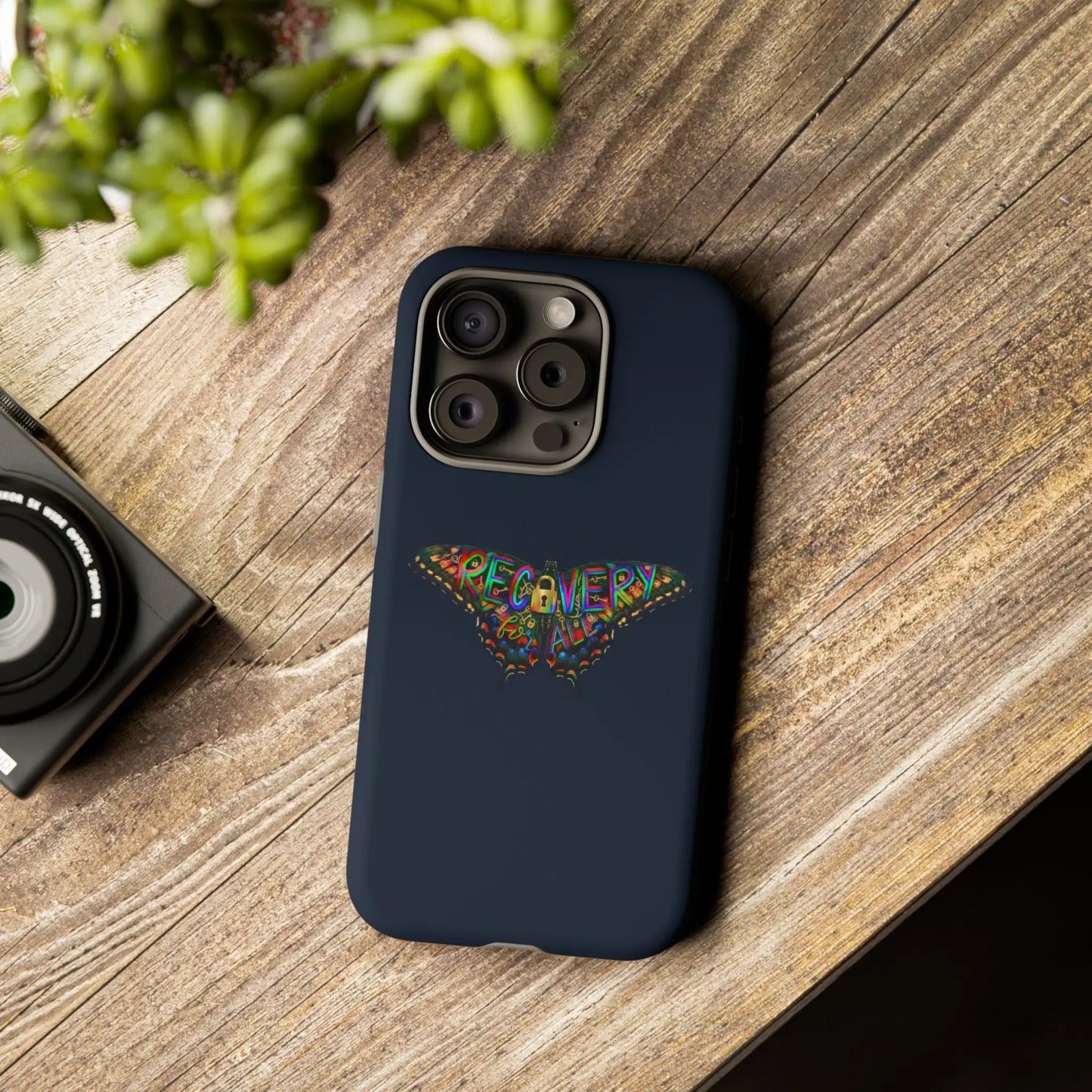 Navy blue phone case with colorful 'RECOVERY' design on a wooden surface with a camera and plant.